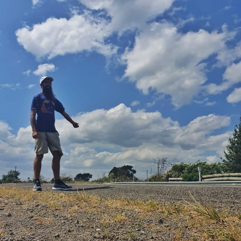 me hitchhiking with thumb out on a partly cloudy day wearing white hat, blue shirt, and shorts.
