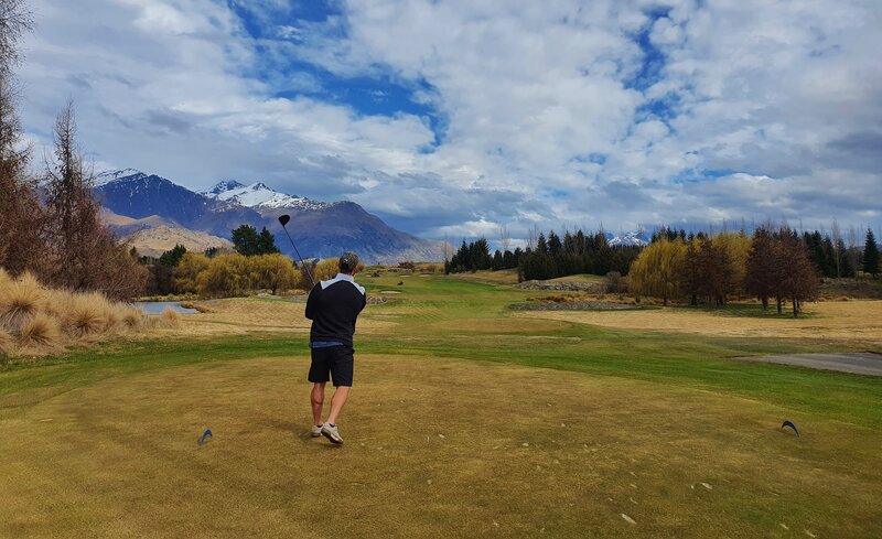 man facing away from the camera having hit a golf shot. golf course looks beautiful with wide open fairway lined with trees on a cloudy day. Mountains with snow caps in the distance.