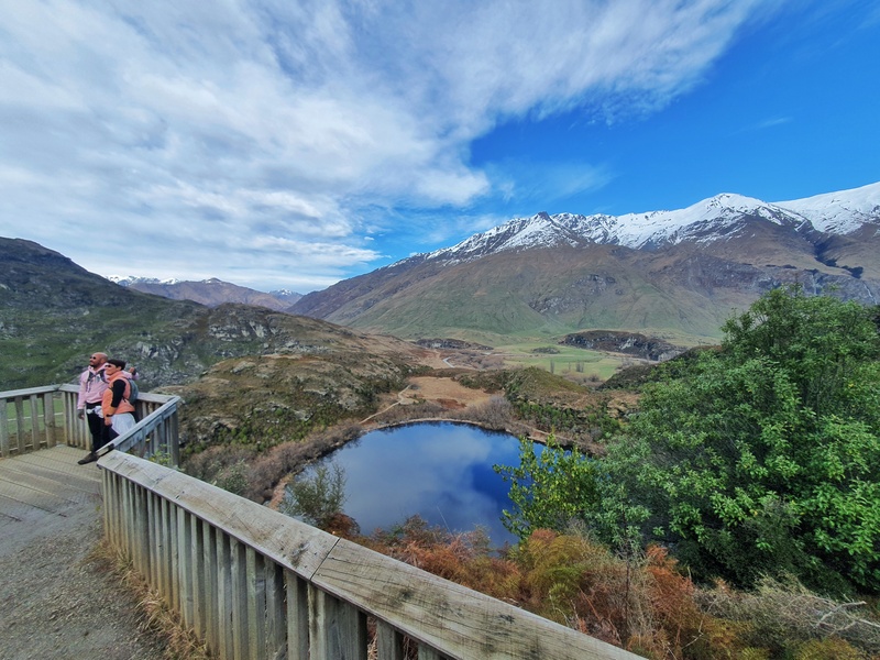 a small lake in the centre of the photo. wooden rail in foreground where two people are seen smiling for a photo. snow capped mountain and cloudy sky in background.