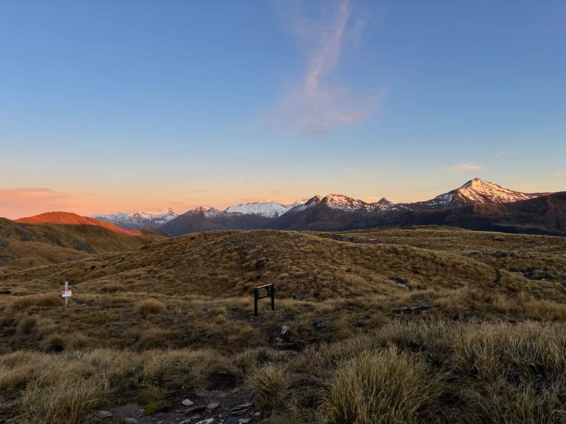 Stunning New Zealand mountain scenery with snow-capped peaks and rolling grassy hills during sunset.