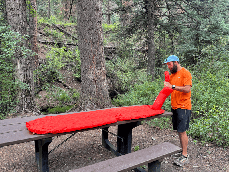 man blowing up a red sleeping mat that's on a picnic bench in the forest